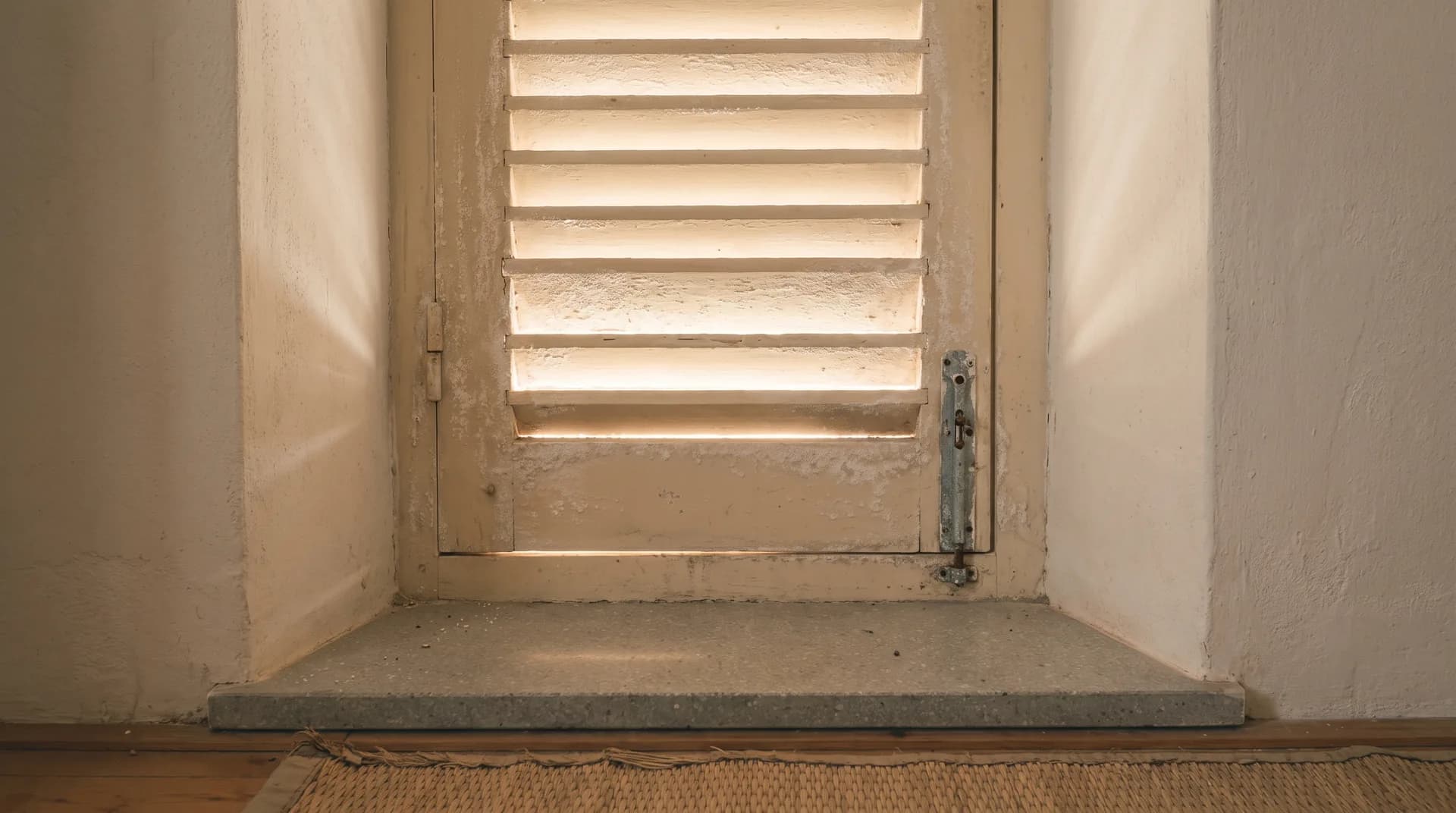 A shuttered window seen from inside a closed house on the Pacific coast, with the salt-hazed light of the dry season falling on the sill and the plaster around it.
