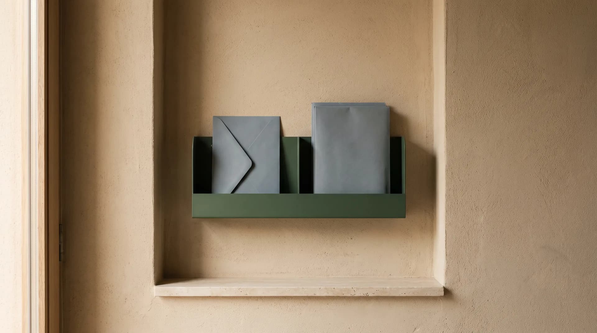 Un nicho de pared enlucido en arcilla color arena con un portacartas de acero verde oscuro y dos sobres lisos sin marcas sobre una repisa de travertino.