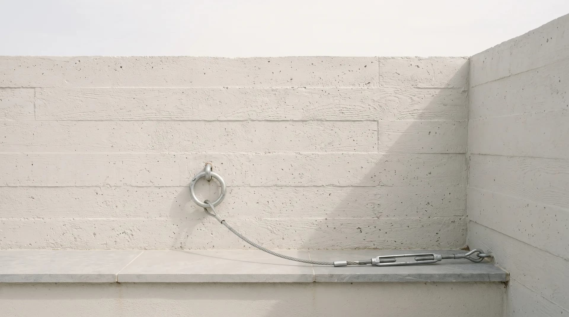 A galvanised steel tie-down ring set into the board-formed concrete of a hillside terrace parapet, with a slack galvanised cable and turnbuckle already fitted to it and nothing yet secured, under flat dry-season daylight.