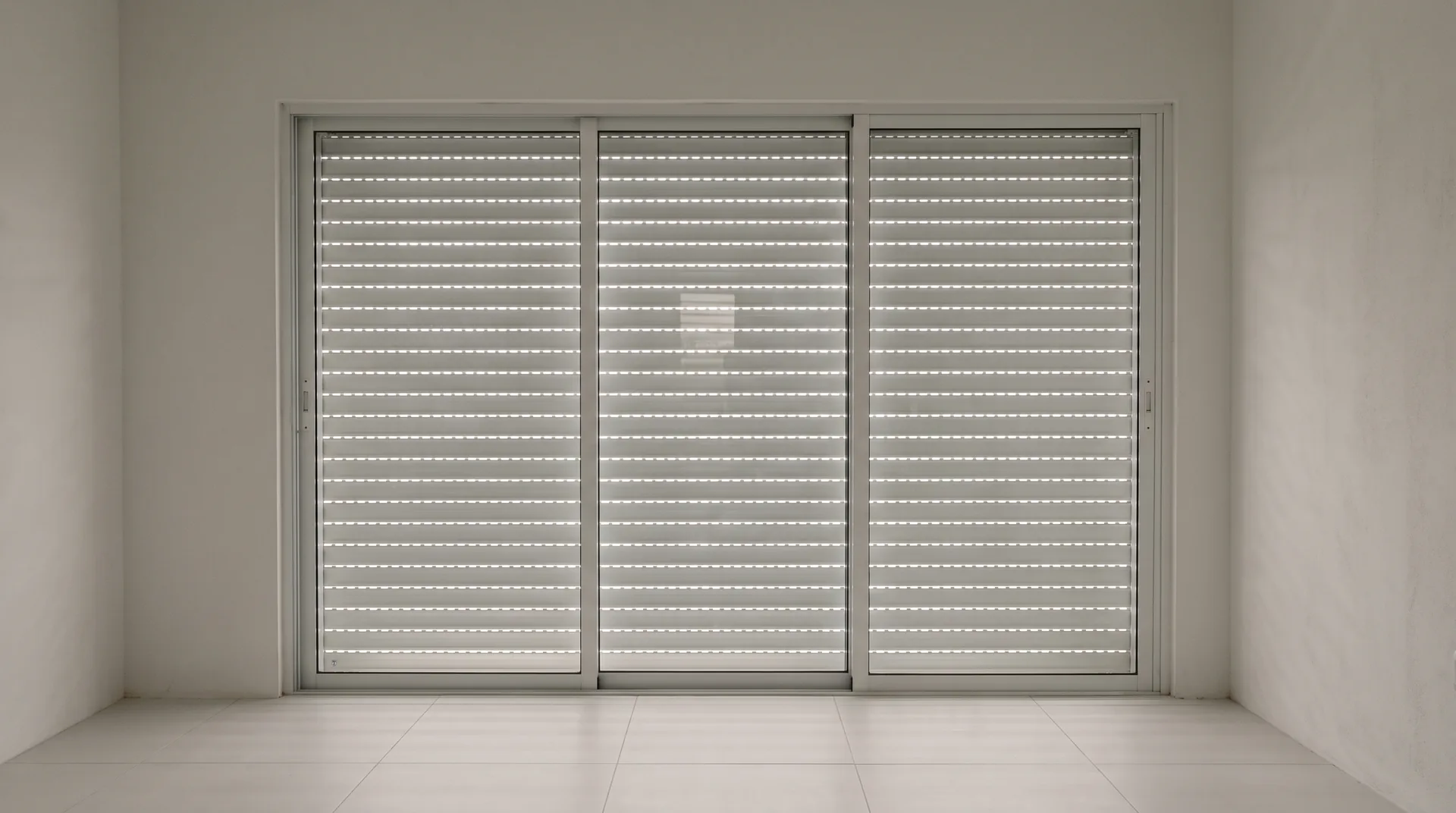 Seen from inside a closed house on the Caribbean coast, a white aluminium accordion storm shutter drawn shut and locked behind a sliding glass door, with thin lines of grey daylight between its slats and a bare porcelain floor in front of it.