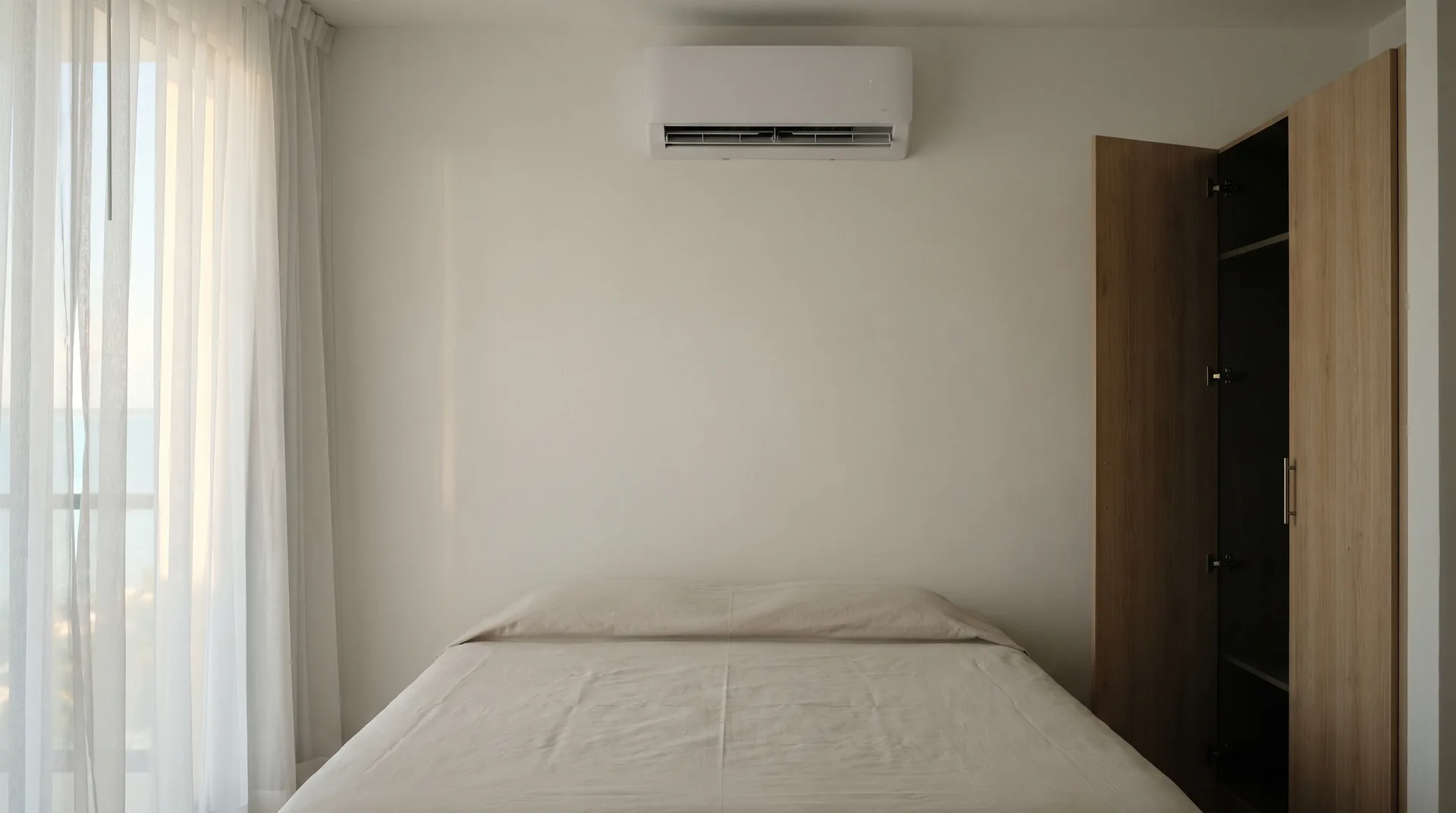 A closed bedroom in a Cancún apartment, with dust covers on the bed, a wardrobe door left ajar for air and a split air conditioner on the wall.