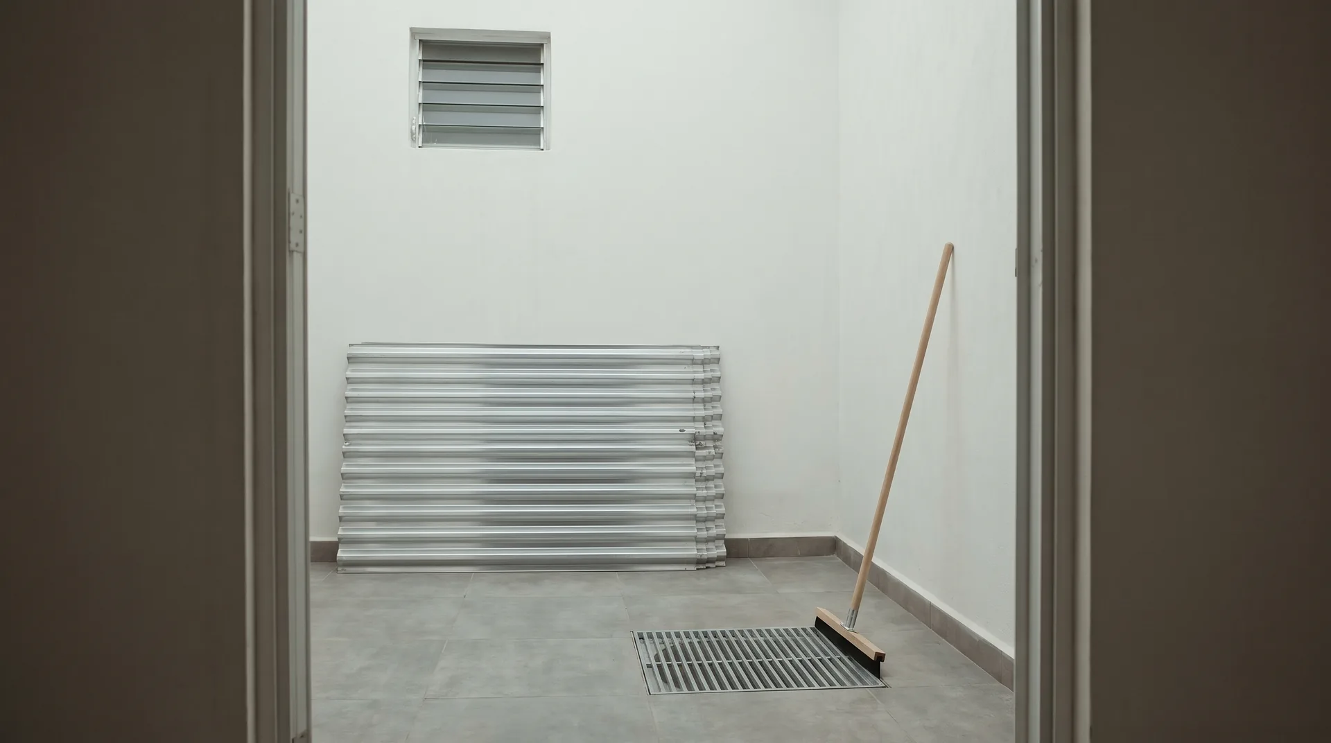The utility room of a closed house in Mexico before the rains, with storm panels stacked against a white wall, a clean floor drain and a squeegee ready.