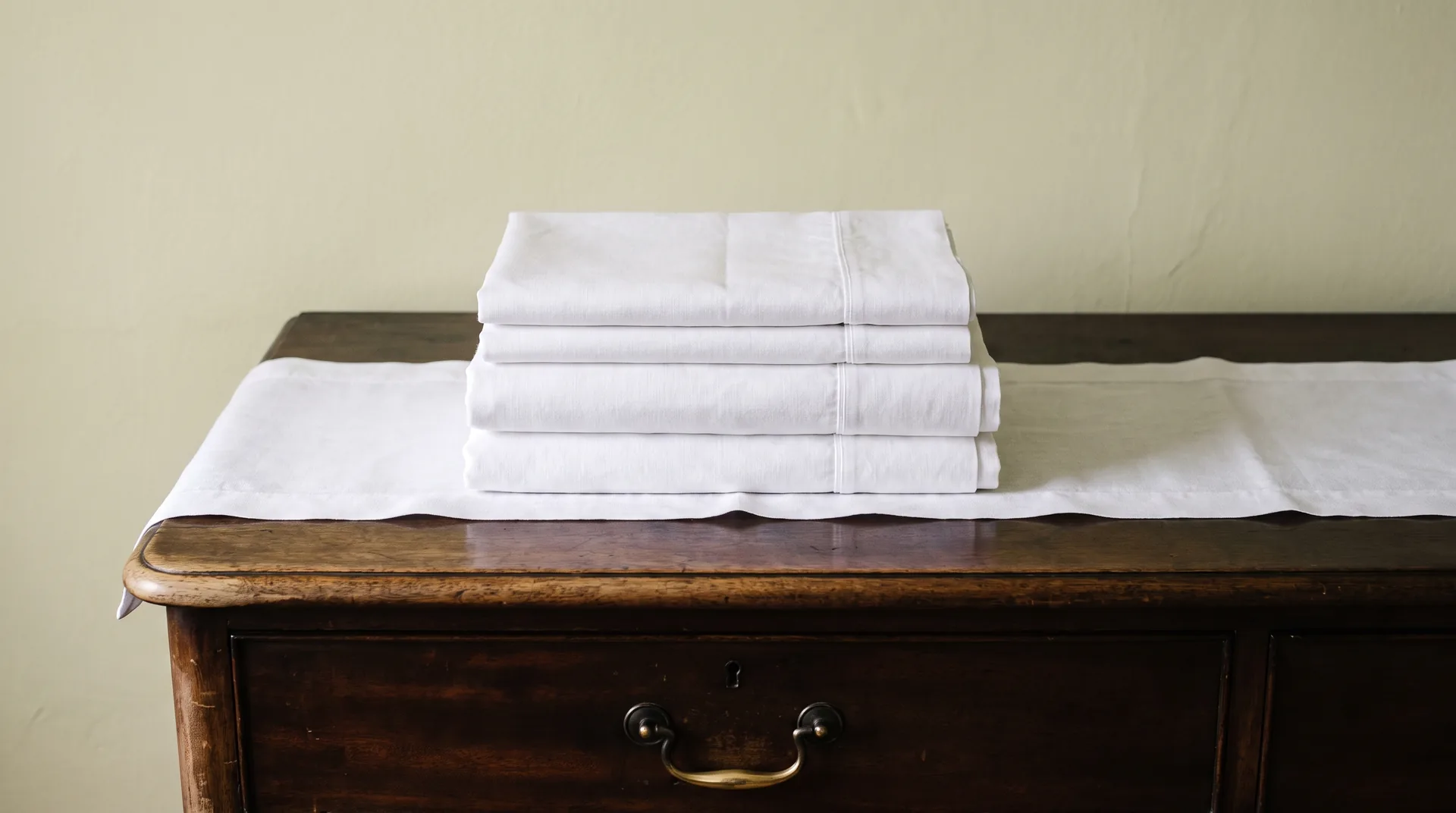 A stack of freshly laundered white linen sheets resting on the waxed top of an old dark chest of drawers, the front edge of the wood worn pale by decades of use.