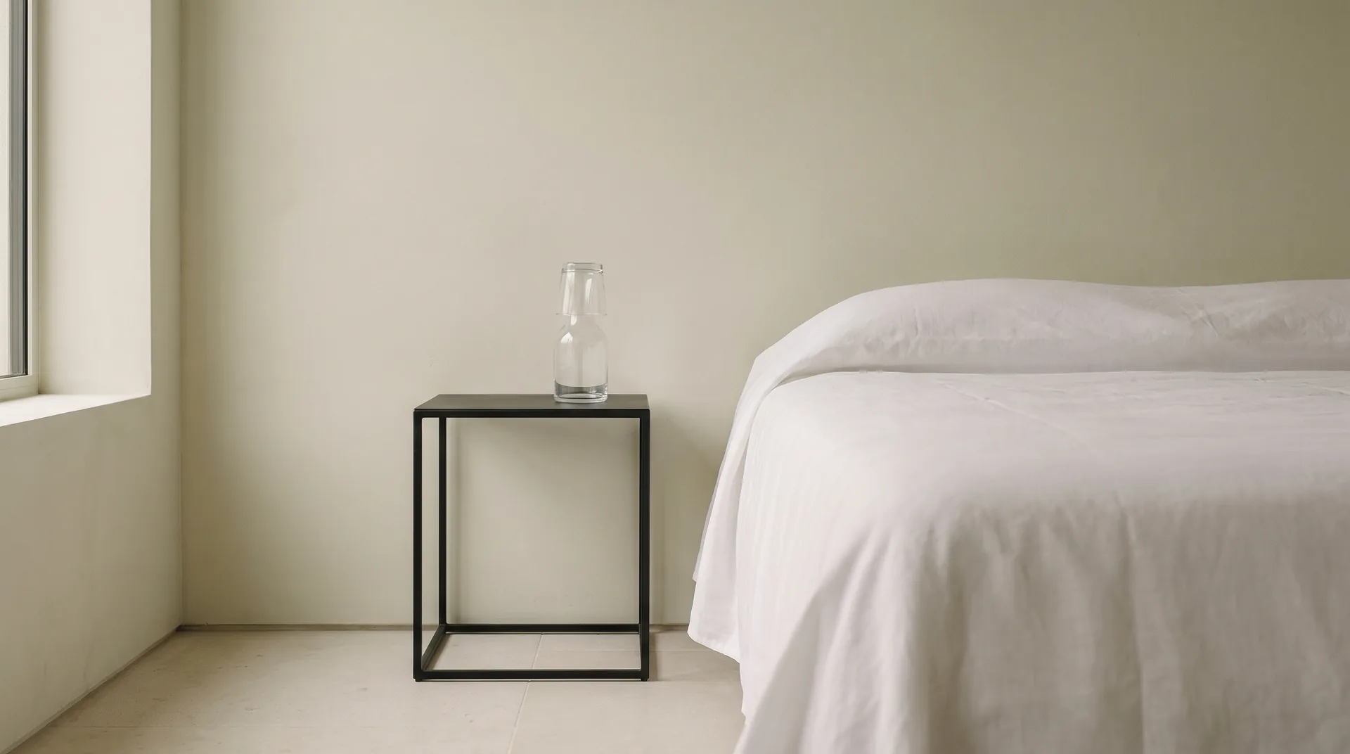 The corner of a guest bedroom prepared for someone who does not live there: a made bed in pressed white cotton, and a plain iron nightstand holding a glass carafe with a tumbler turned upside down over its neck, and nothing else at all.